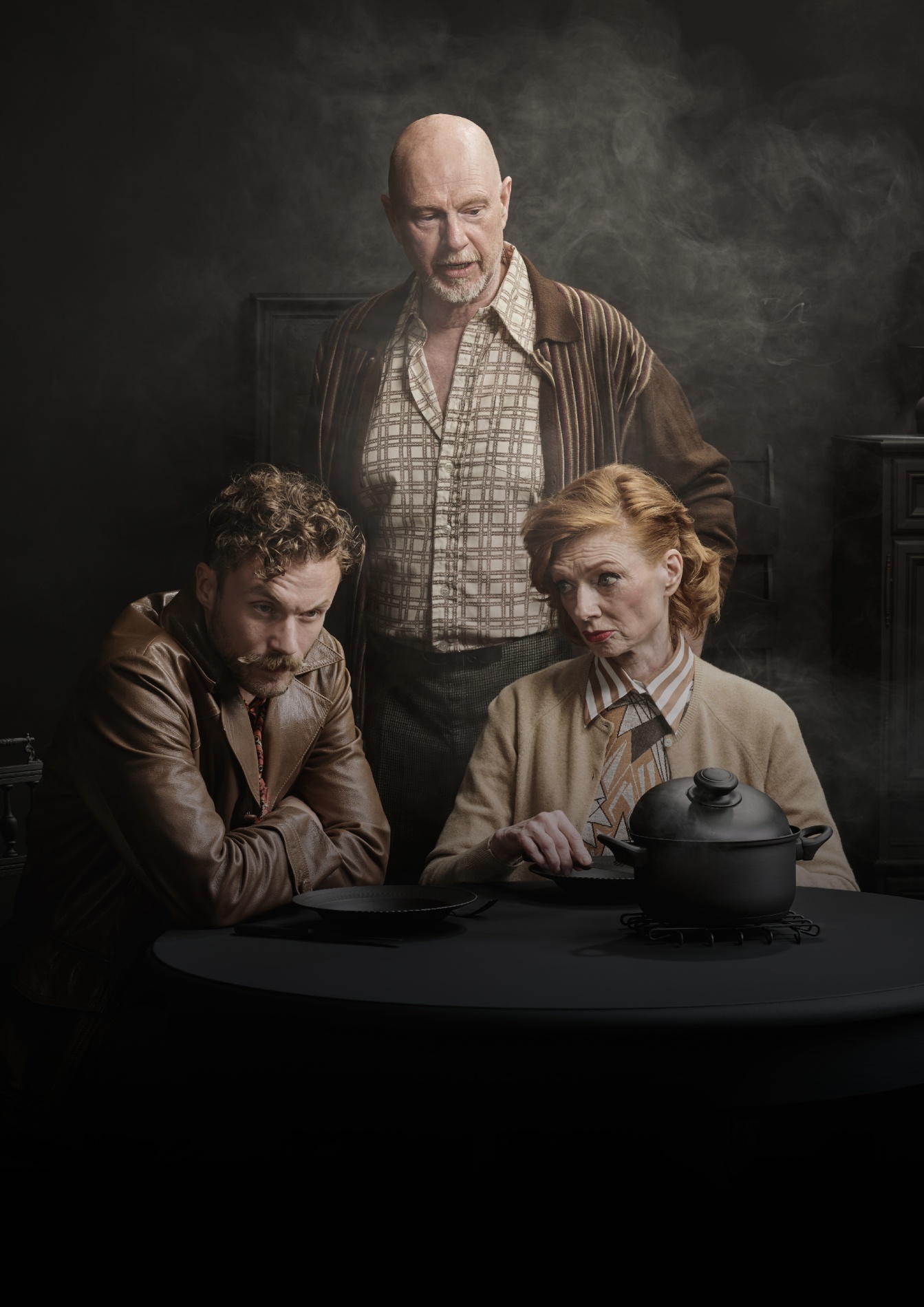 Mensen zitten aan een tafel, met een zwarte achtergrond en een waas van rook. De vrouw links heeft rood haar en kijkt sceptisch naar de man rechts, die een lepel vasthoudt. De man links heeft krullend haar en een baard en kijkt serieus naar de tafel. De man achter staat en kijkt bezorgd of nadenkend naar de groep.
