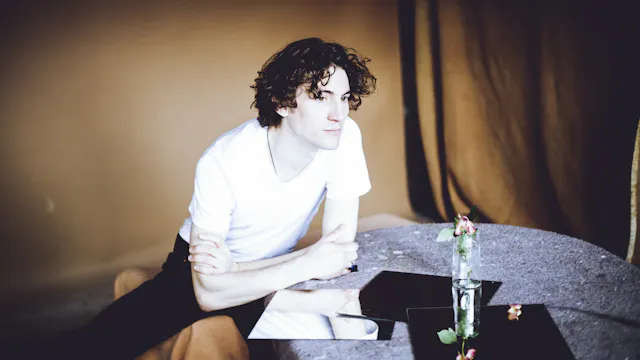 Jongeman in wit T-shirt leunend op een tafel met spiegels en bloemen.