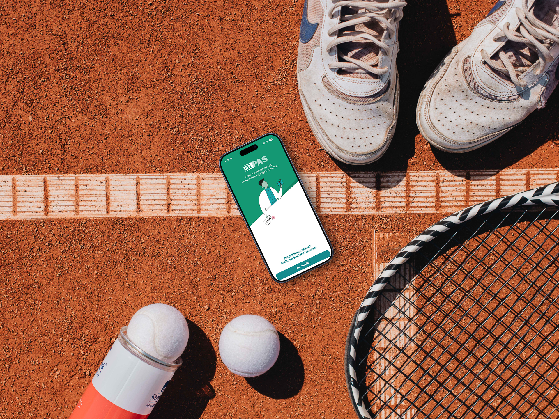 Bovenaanzicht van een stuk gravel tennisveld met racket, ballen, sportschoenen en een smartphone waarop het aanmeldscherm van de UiTPAS-app te zien is