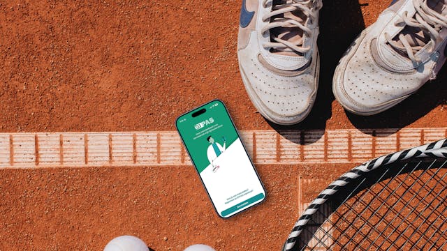 Bovenaanzicht van een stuk gravel tennisveld met racket, ballen, sportschoenen en een smartphone waarop het aanmeldscherm van de UiTPAS-app te zien is