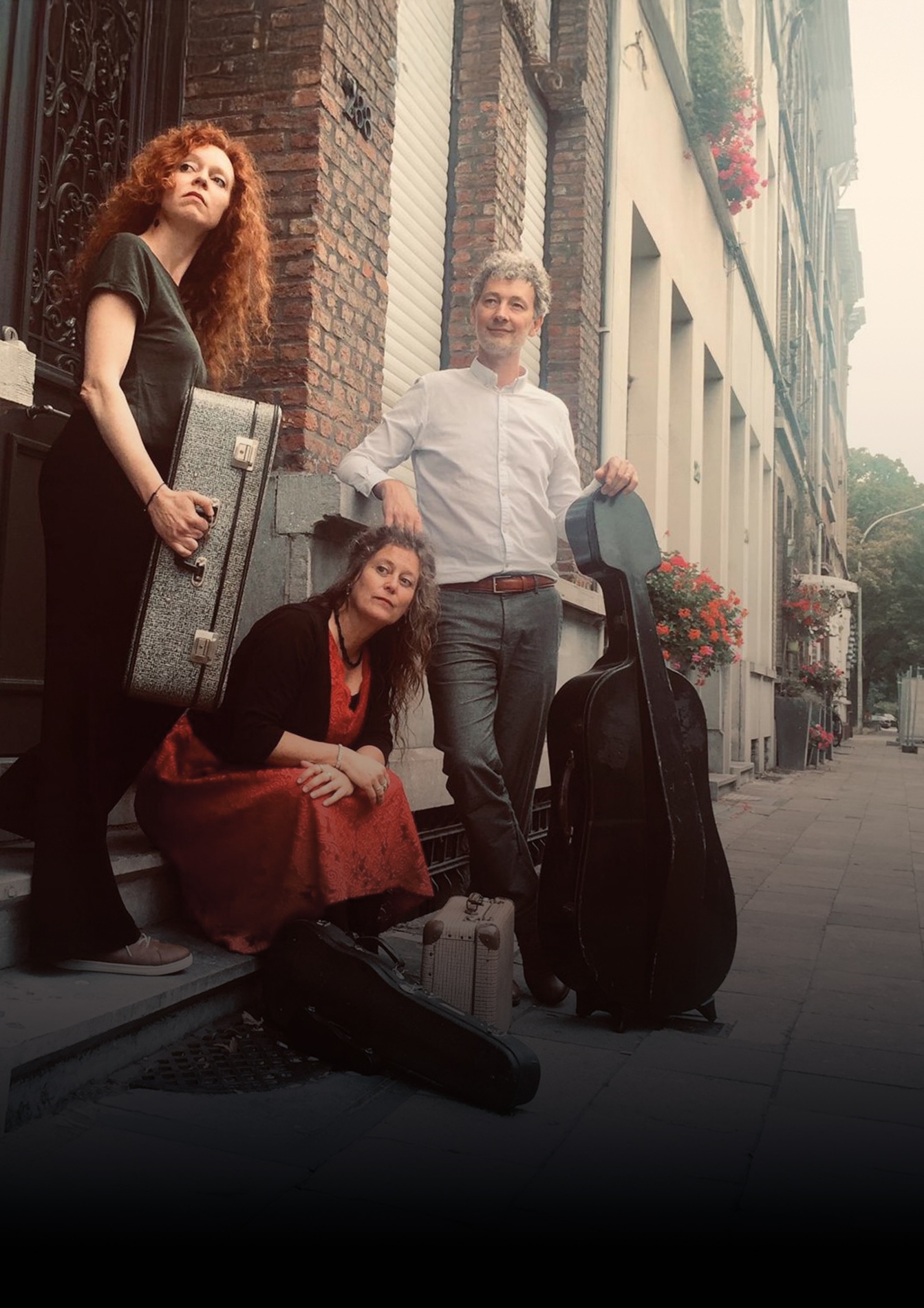 Drie mensen met muziekinstrumenten staan op een stoep voor een gebouw met bakstenen en bloemenbakken. Eén persoon staat met een gitaar, een ander zit op de treden met een viool en een derde persoon staat met een koffer en een gitaar.