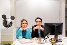 Afbeelding van 2 personen die aan een bureau zitten en naar de camera te kijken.