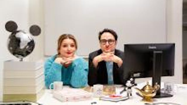 Afbeelding van 2 personen die aan een bureau zitten en naar de camera te kijken.