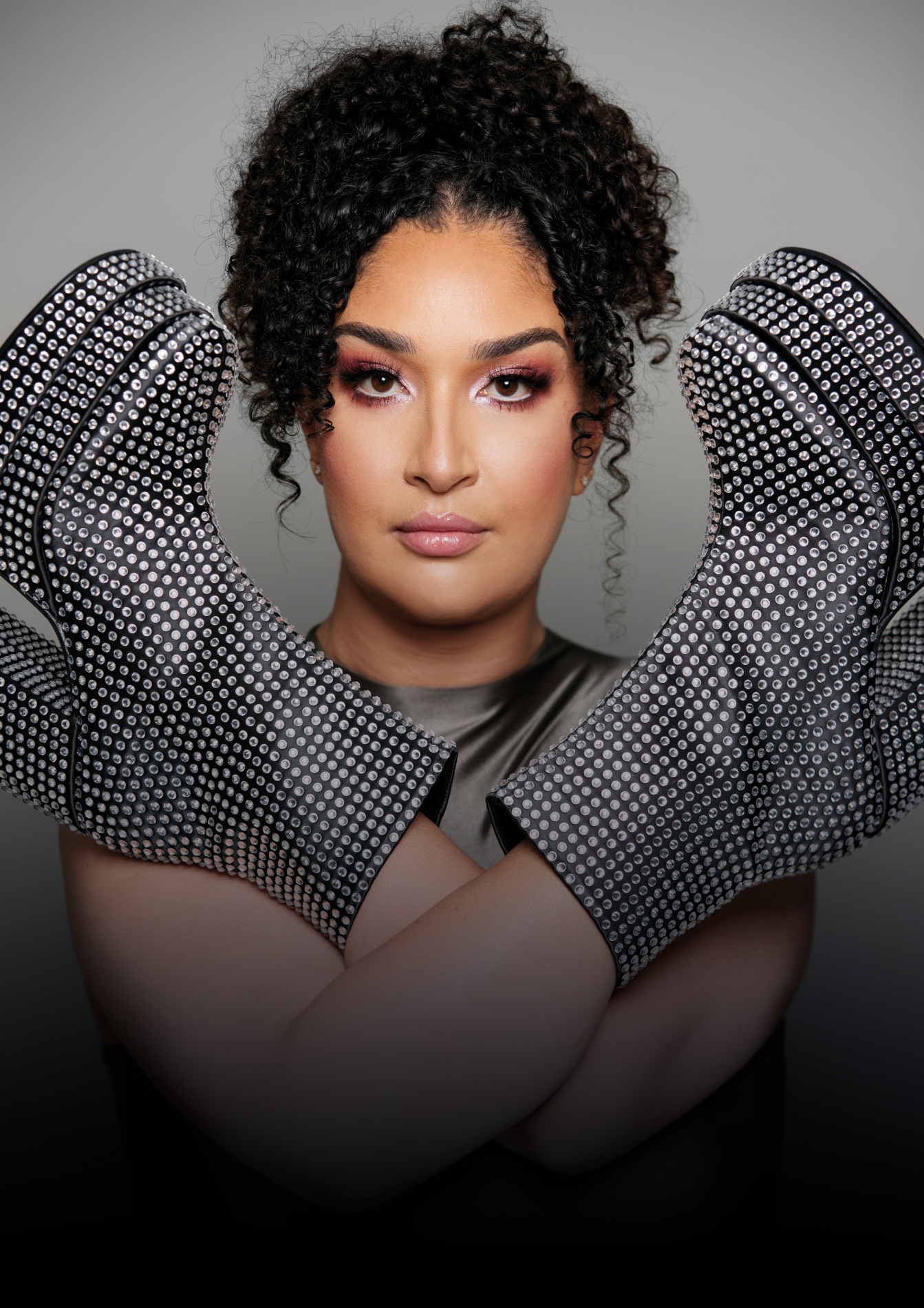 Een vrouw met gekruld haar en make-up kijkt recht in de camera. Ze draagt opvallende handschoenen bedekt met glinsterende steentjes en heeft haar armen gekruist voor haar borst. De achtergrond is grijs.