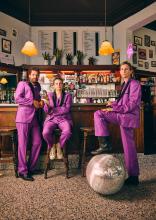 Drie personen in een magenta pak voor een bar toog. 1 van de personen heeft een voet op een discobal dat op de grond ligt.