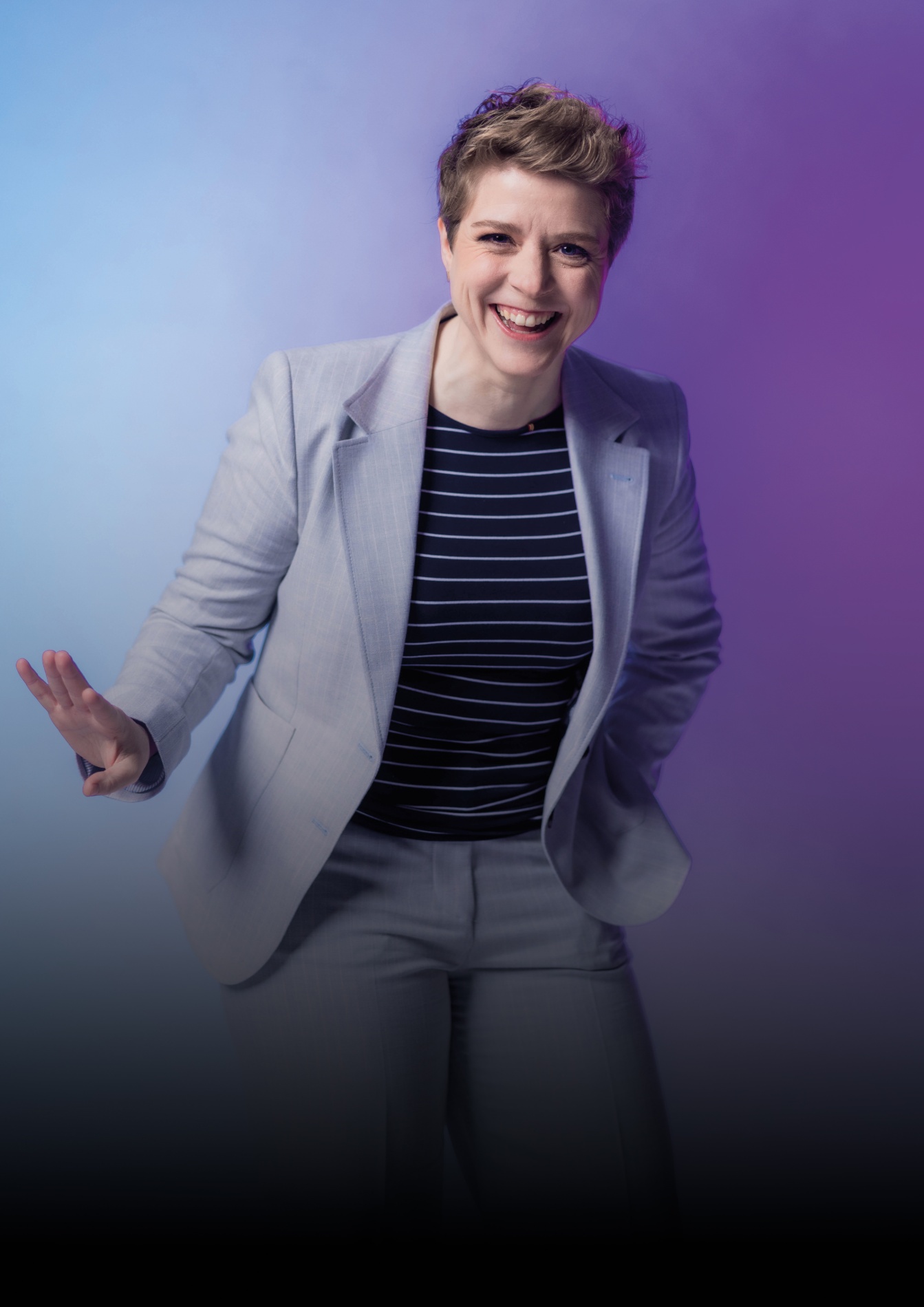Een vrouw met kort, lichtbruin haar die glimlacht en een handgebaar maakt, gekleed in een lichtgrijze blazer en een zwart-wit gestreepte top, staand voor een achtergrond met een verloop van blauw naar paars.