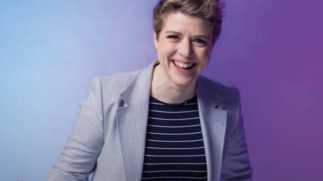 Een vrouw met kort, lichtbruin haar die glimlacht en een handgebaar maakt, gekleed in een lichtgrijze blazer en een zwart-wit gestreepte top, staand voor een achtergrond met een verloop van blauw naar paars.