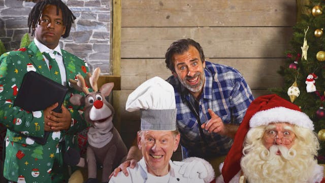 Een groep mensen in een feestelijke setting met kerstdecoraties. Er is een man gekleed als kerstman, een chef-kok met een schotel vol kerstballen, een man met een blauwe geruite overhemd die glimlacht, een jongen in een groen kerstpak, en een poppenversie van een rendier. Ze poseren allemaal vrolijk voor de camera, met een kerstboom en een kerstkrans op de achtergrond.