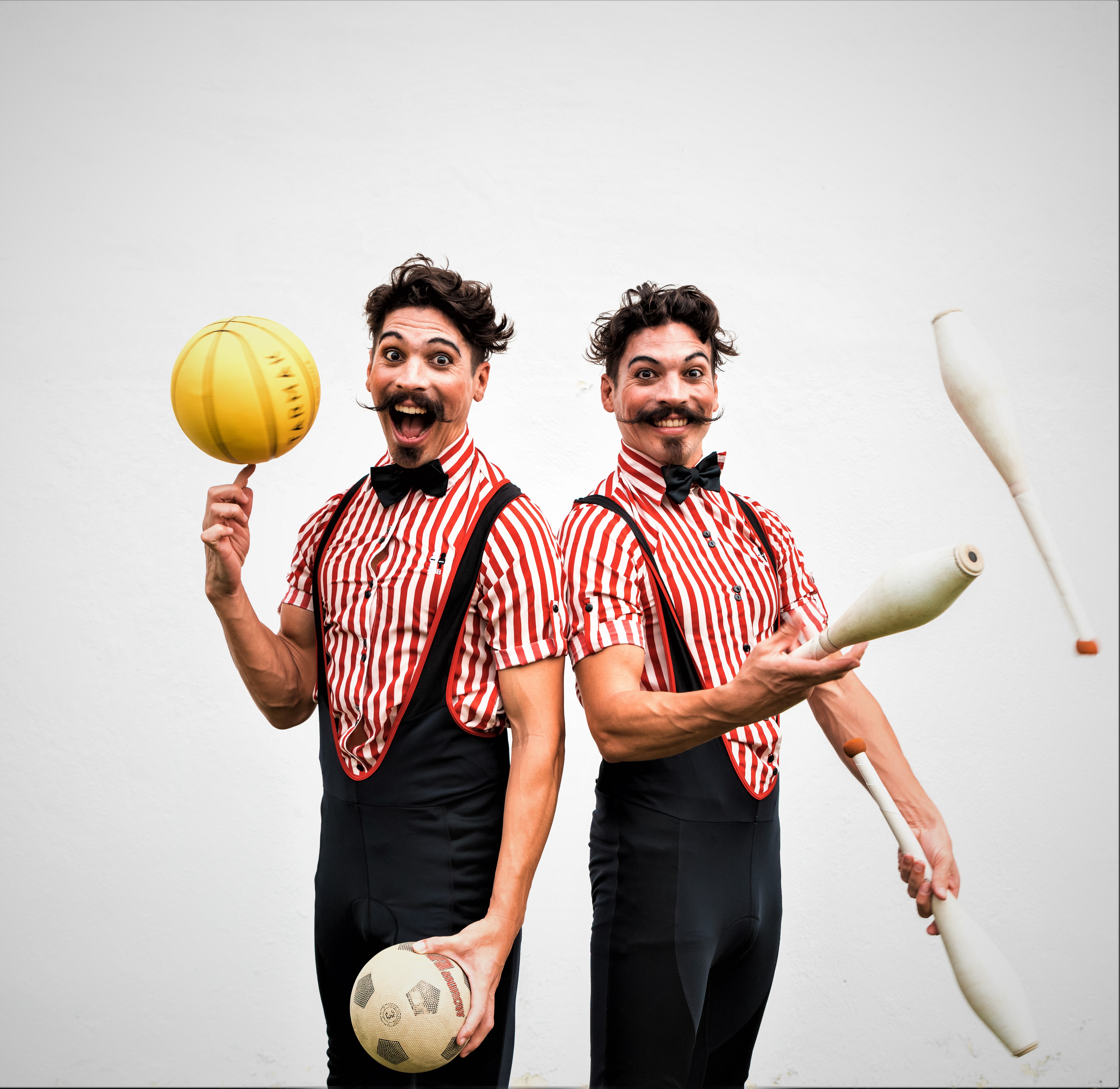 Twee vrolijke artiesten trekken gekke gezichten met kegels en ballen in hun handen. Ze dragen beiden een rood-wit gestreept hemd en een bruine salopet.