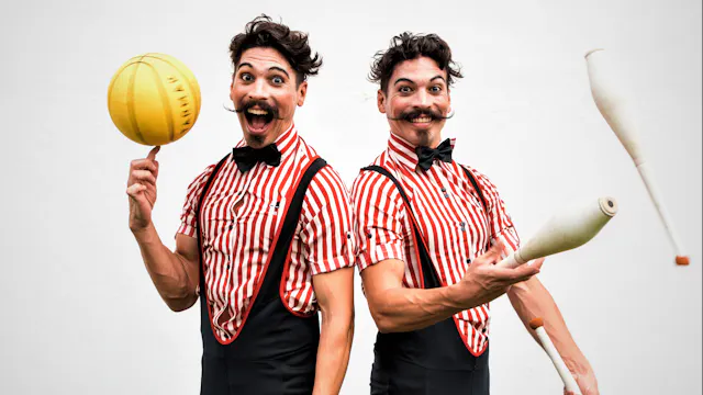 Twee vrolijke artiesten trekken gekke gezichten met kegels en ballen in hun handen. Ze dragen beiden een rood-wit gestreept hemd en een bruine salopet.