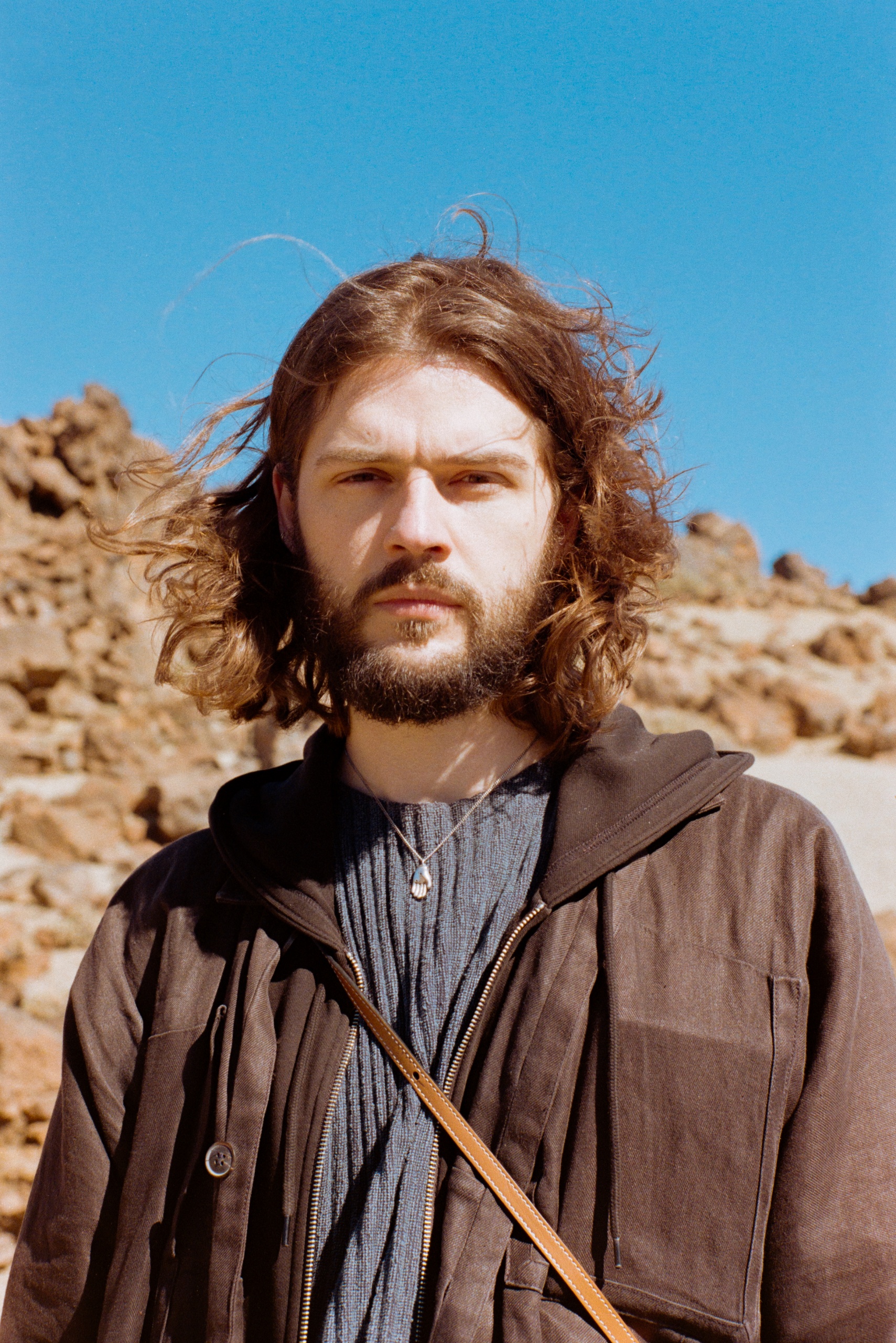 Jongeman met een baard en krullend haar, staand in een woestijnachtige omgeving.
