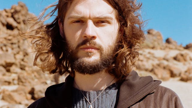 Jongeman met een baard en krullend haar, staand in een woestijnachtige omgeving.