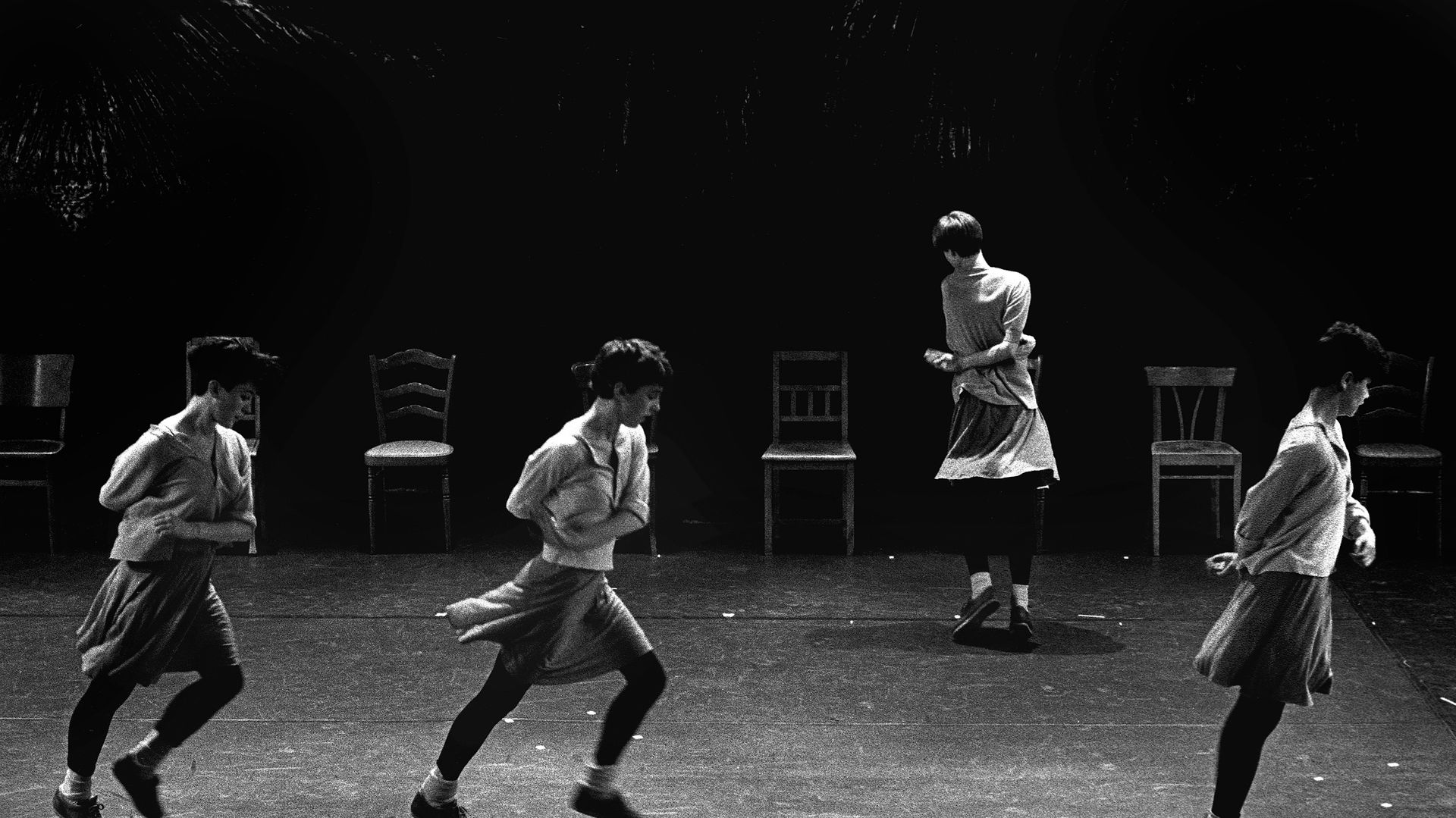 Zwart-wit scènebeeld uit Rosas danst Rosas met vier danseressen en vier stoelen.