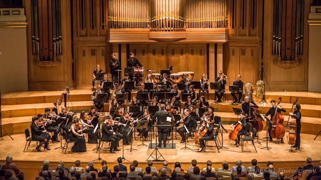 Een orkest speelt op een podium met een grote orgel achtergrond.