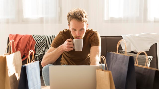 man omringd door winkeltassen, die kijkt op laptopscherm terwijl hij koffie drinkt