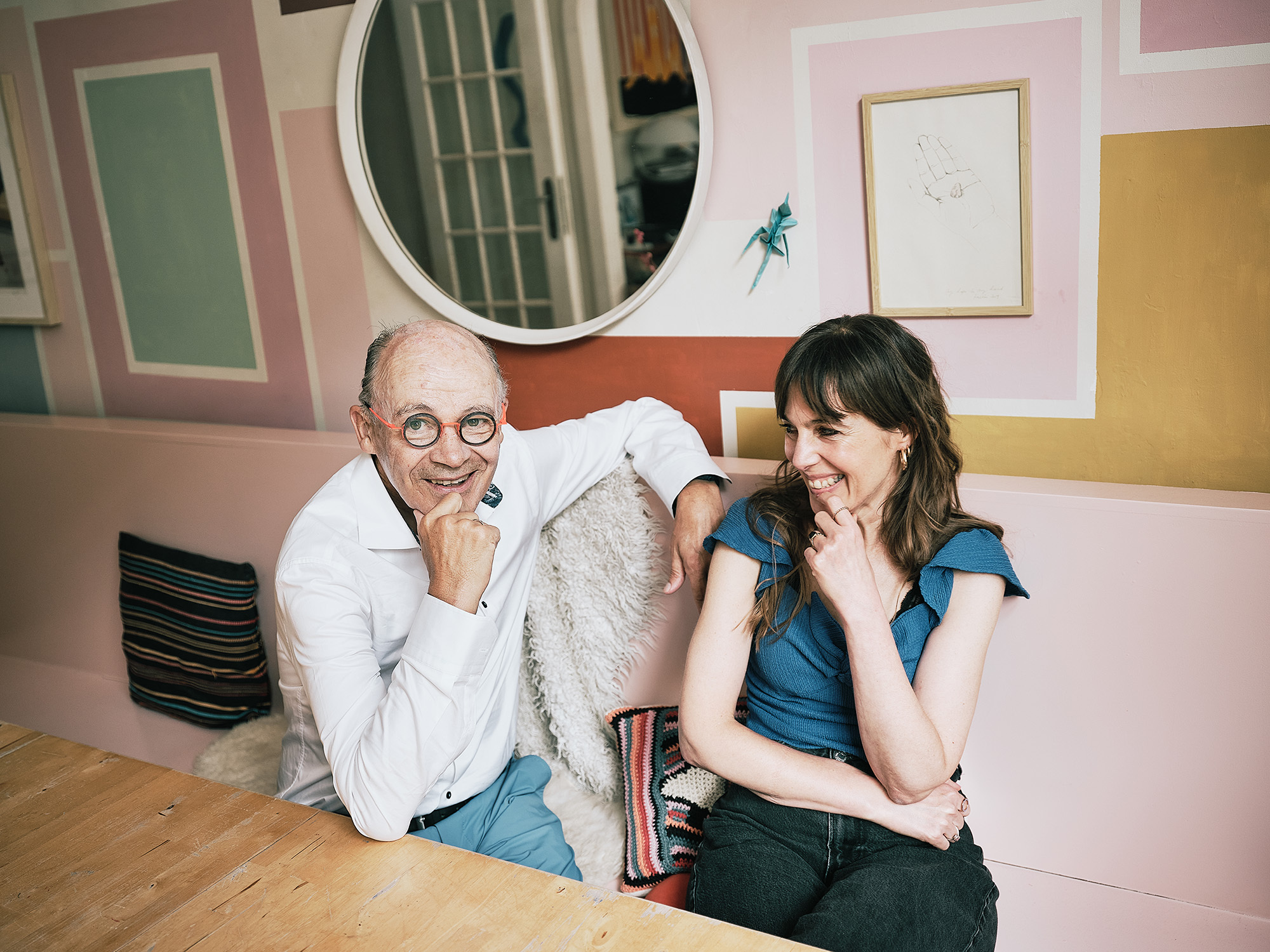Jeugdpsychiater Peter Adriaenssens en kunstenares Kristien Dirkx zitten aan een houten tafel; achter hen hangen er kleurrijke kunstwerken aan de muur