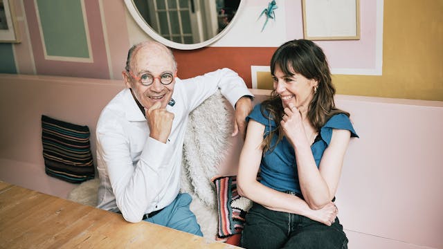 Jeugdpsychiater Peter Adriaenssens en kunstenares Kristien Dirkx zitten aan een houten tafel; achter hen hangen er kleurrijke kunstwerken aan de muur