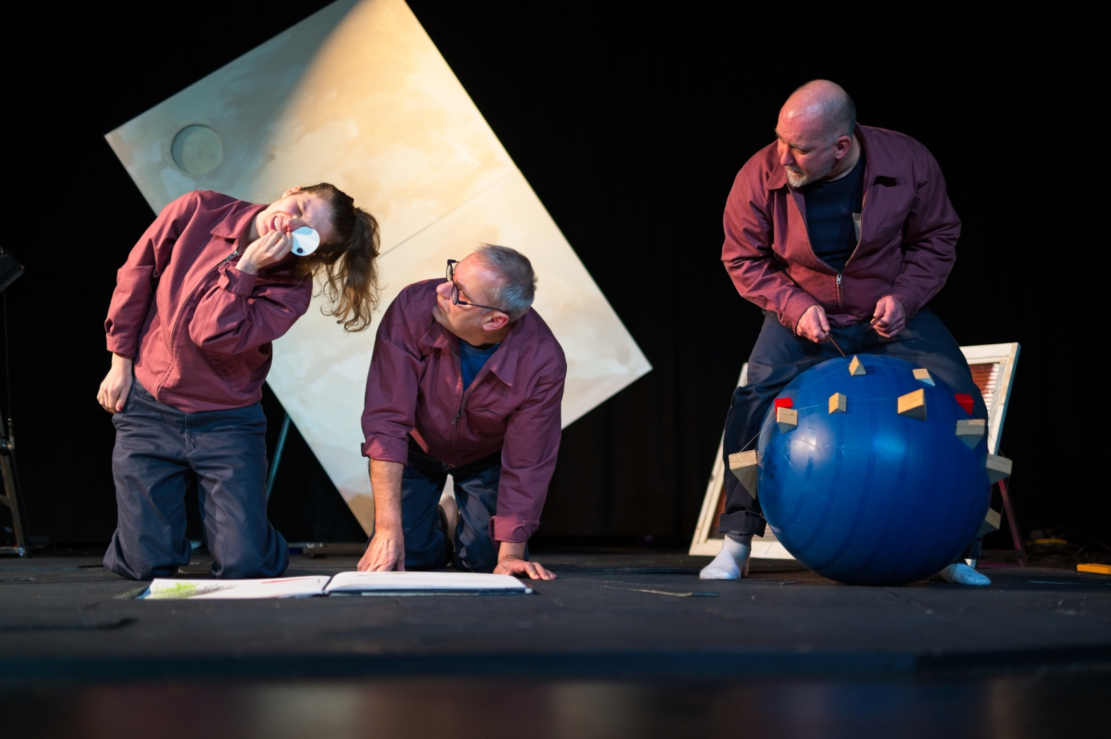 Drie acteurs op een podium met props, in een theatrale scène.