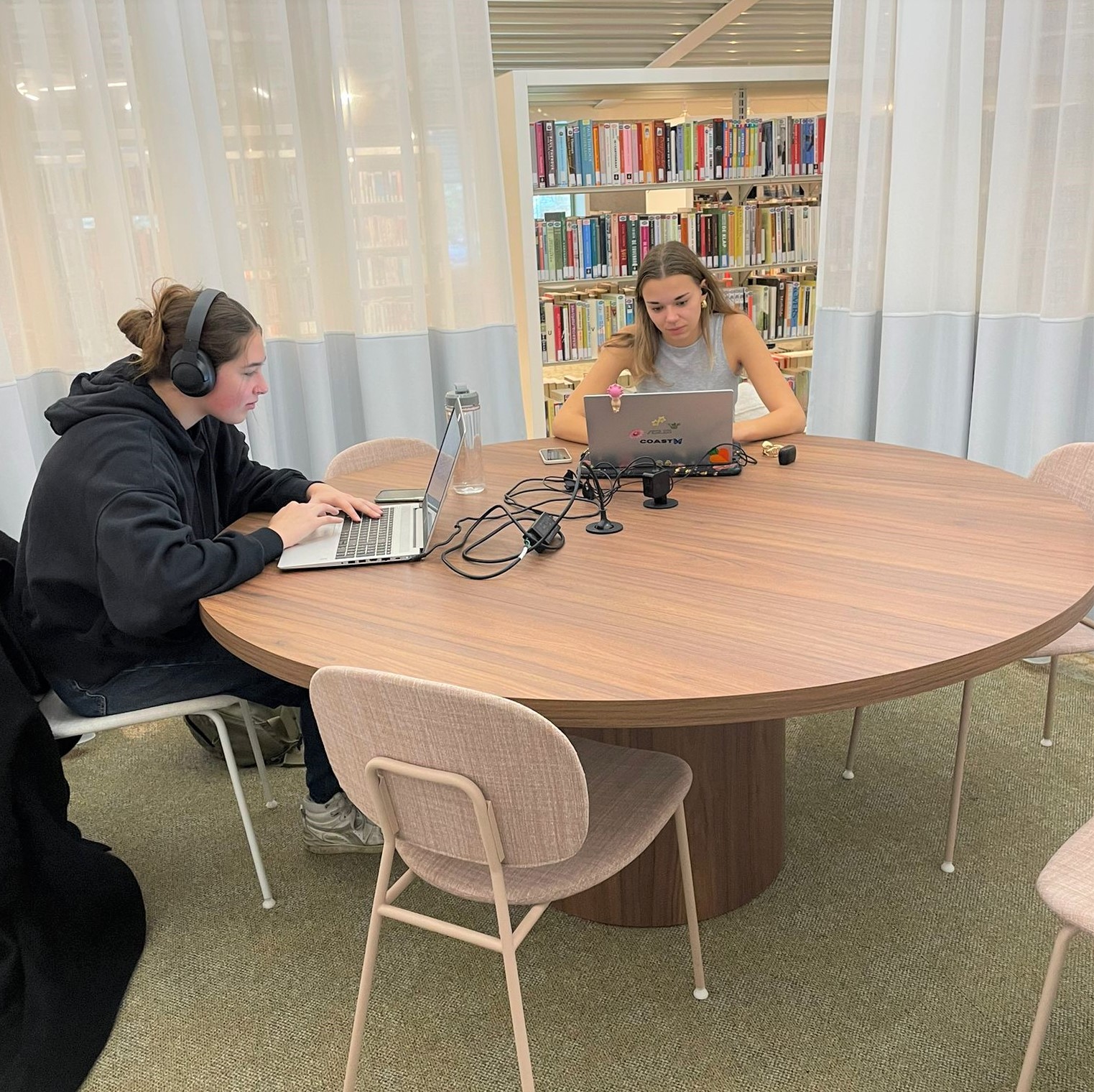 Twee jonge vrouwen werken geconcentreerd op laptops aan een ronde tafel in een bibliotheek.